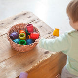 Vor einem Kleinkind steht ein Weidenkorb mit den Grapat "Mates" Töpfchen aus Holz in allen 12 Regenbogenfarben.