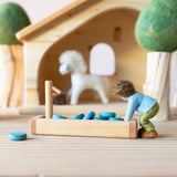 Kleine blaue Mandalasteine liegen in einer Holztränke zum Spielen, eine kleine Filzfigur beugt sich zur Tränke.