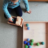 Ein Kind setzt Filzbälle von Kokomoo auf die Ladefläche vom Fagus Unimog, daneben ein großes Holztablett mit weiteren Filzkugeln in Blau- und Grüntönen.