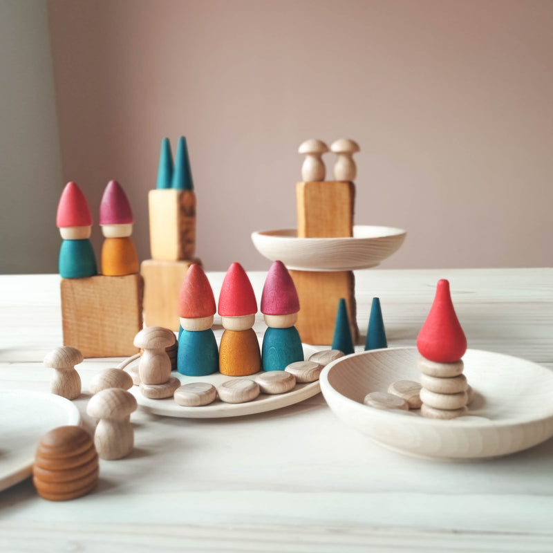 Eine Spielsituation mit bunten, rot-grün-gelben Holzzwergen von Grapat. Sie stehen auf und in Holzschalen und Holztellern. Einige Bausteine stehen in der Landschaft. Auf ihnen stehen kleine grüne Kegel und Minipilze aus Holz.