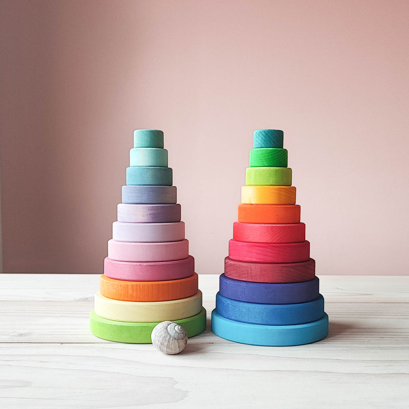 Ein blick von vorne auf 2 Stapeltuerme aus bunt lasiertem Holz der firma Grimms. llinks sieht man den Turm in Pastellfarben (von guen,ueber gelb, orange, Rosatoene bis helltuerkis. Daneben steht der kraetig bunte Stapelturm. vor dem linken Turm liegt ein schneckenhaus einer Weibergschnecke, damit man das Groessenverhaeltnis besser erkennen kann.