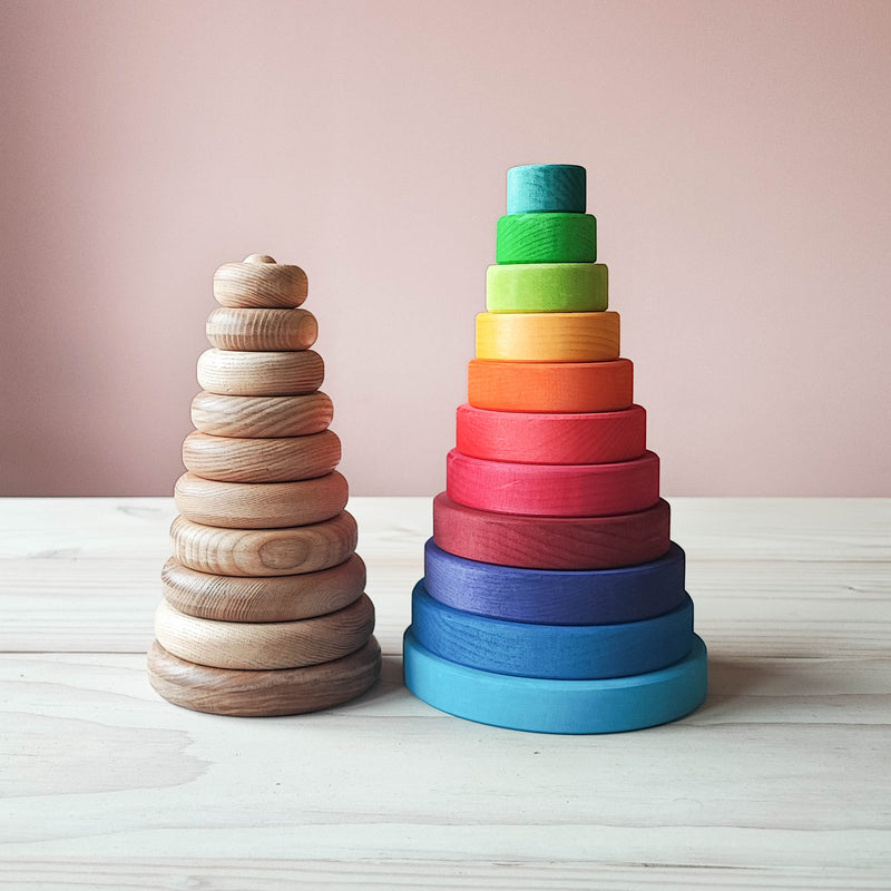 Ein Blick von vorne auf 2 verschiedene Stapeltuerme. Der linke hat runde Naturholzscheiben - ohne Farbe.Die einzelnen Ringe 10 an der zahl sind zwar flach gearbeitet, aber die Kanten geschmeidig abgerundet.  Daneben steht der Scheibenturm von Grimms in Regenbogenfarben. Dieser ist ein Stueck hoeher, hat etwas mehr Durchmesser und die buntlasierten Scheiben sind an den Aussenkanten eher kantig, als abgerundet.
