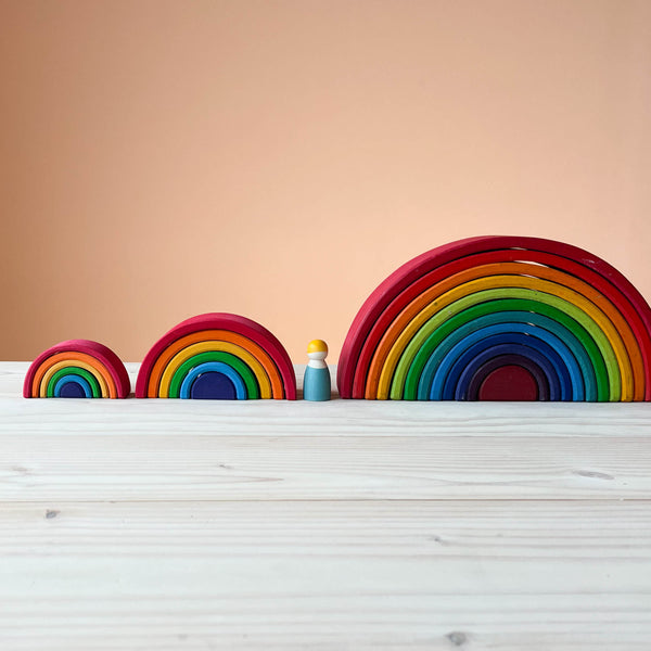 Alle drei Regenbogen von Grimms in den klassischen sechs Regenbogenfarben nebeneinander aufgestellt, ganz links der Mini Regenbogen, in der Mitte der kleine und ganz rechts der große Regenbogen. Eine Holzfigur steht auch dazwischen.