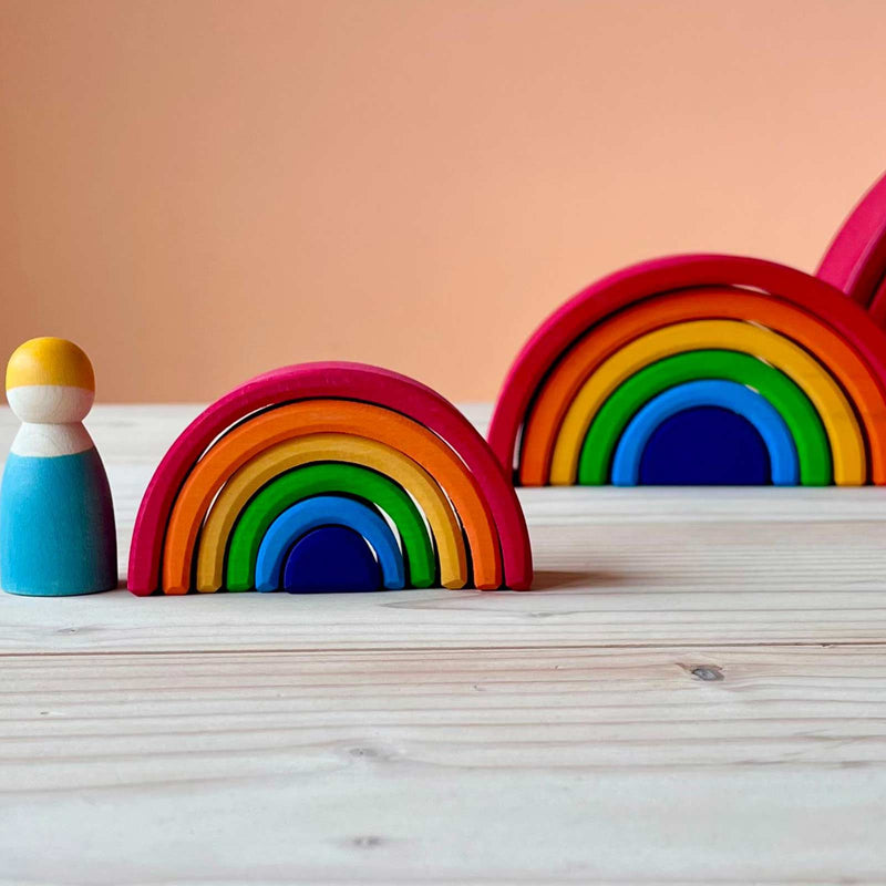 Der Regenbogen Mini von Grimms links daneben steht eine kleine Spielfigur aus Holz, im Hintergrund ist noch der kleine Regenbogen und noch ein bisschen vom großen Grimms Regenbogen am rechten Rand zu erkennen.