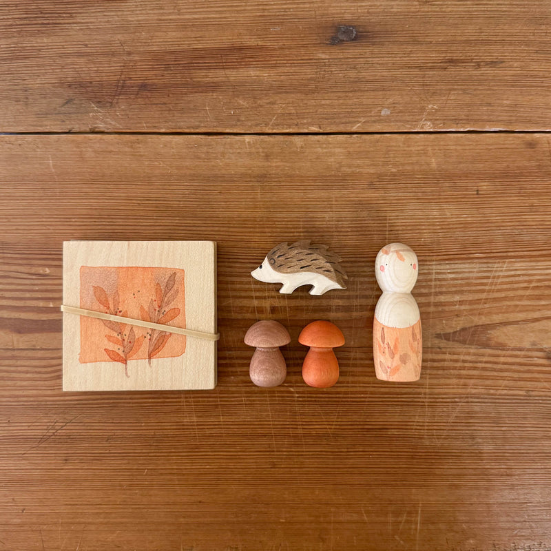 Das Spiele- und Forscherset für Kinder "Hello Autumn" von Grapat und Ostheimer liegt auf einem Tisch. Der Igel, die beiden Holzpilze, die Nins Spielfigrt und die kleine Blätterpresse.