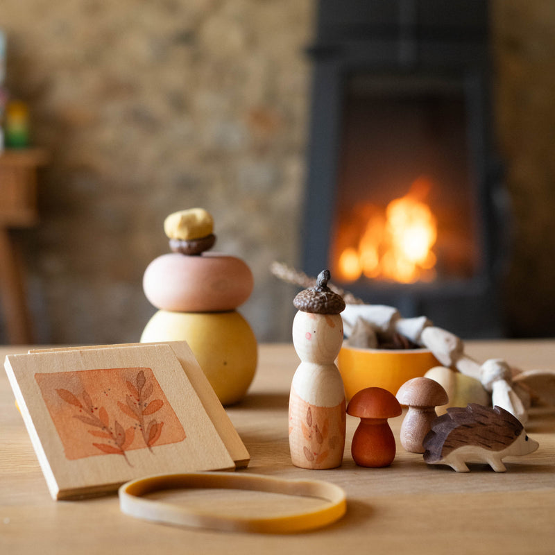 Das Spiele- und Forscherset für Kinder "Hello Autumn" von Grapat und Ostheimer steht auf einem Tisch vor dem Kamin. Der Igel, die beiden Holzpilze, die Nins Spielfigur und die kleine Blätterpresse.
