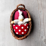 Das Wichtelnest von Kokomoo in rot: ein Weidenkörbchen als Puppenbett, in dem ein weißer Nanchen Wichtel in einer rot gemusterten Bettwäsche kuschelt. 