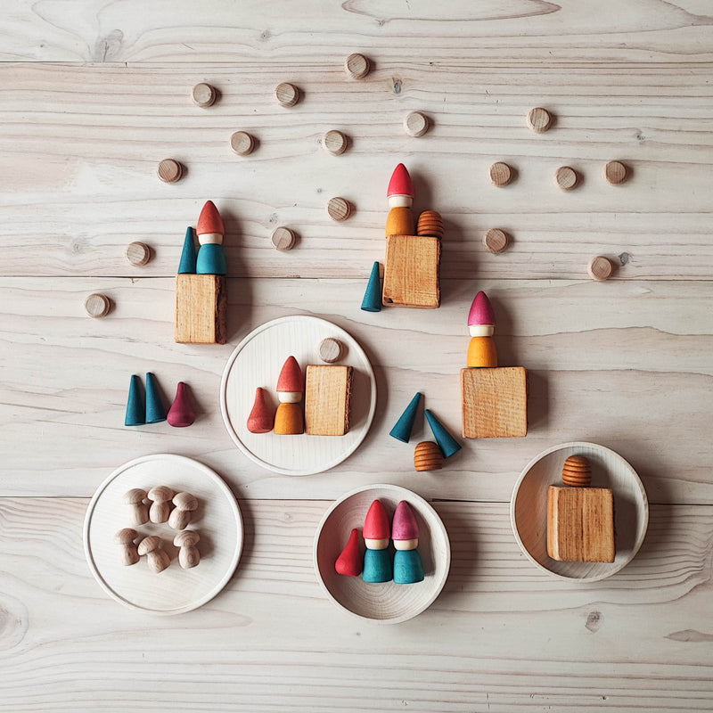 6 rot-grüne Holzzwerge von Grapat auf runden kleinen Holztellern und in Holzschalen. Um sie herum verteilt liegen kleine runde Holzplättchen, kleine grüne Kegel und Mini-Holzpilze.