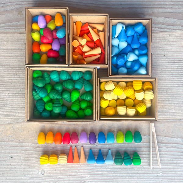 Ein Blick von oben auf 5 Pappkartons, darin befinden sich kunterbunte kleine Mandala-Holzmaterialien in verschiedennen Farben - nach Formen sortiert. Bunte Regenbogeneier, kleine orangene Kegel, Tropfen in 3 Blautönen, Kleine grüne Bäume, die wie Tannenwipfel aussehen und gelbe Honigwaben. Davor liegen von jeder Form und Farbe jeweils 1Stück aufgereiht. Daneben liegt eine kleine Mini-Holzzange, wie eine Gurkenzange, nur klein.