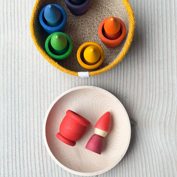 Das Kokomoo Bunte Zwerge Spielset für Babys ab 1 Jahr, Eine rote Holzfigur und ein kleines Töpfchen aus Holz liegen in der Holzschale, daneben der Jutekorb mit den restlichen Grapat Mates und Grapat Tomtens in Regenbogenfarben.