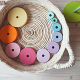 Zehn Scheiben des Grimms Scheibenturm in Pastell liegen kreisförmig am Rand in einem hellen flachen Korb. die Scheiben beginnen links oben mit der groessten in pastellgelb, und werden bis rechts oben immer kleiner. 