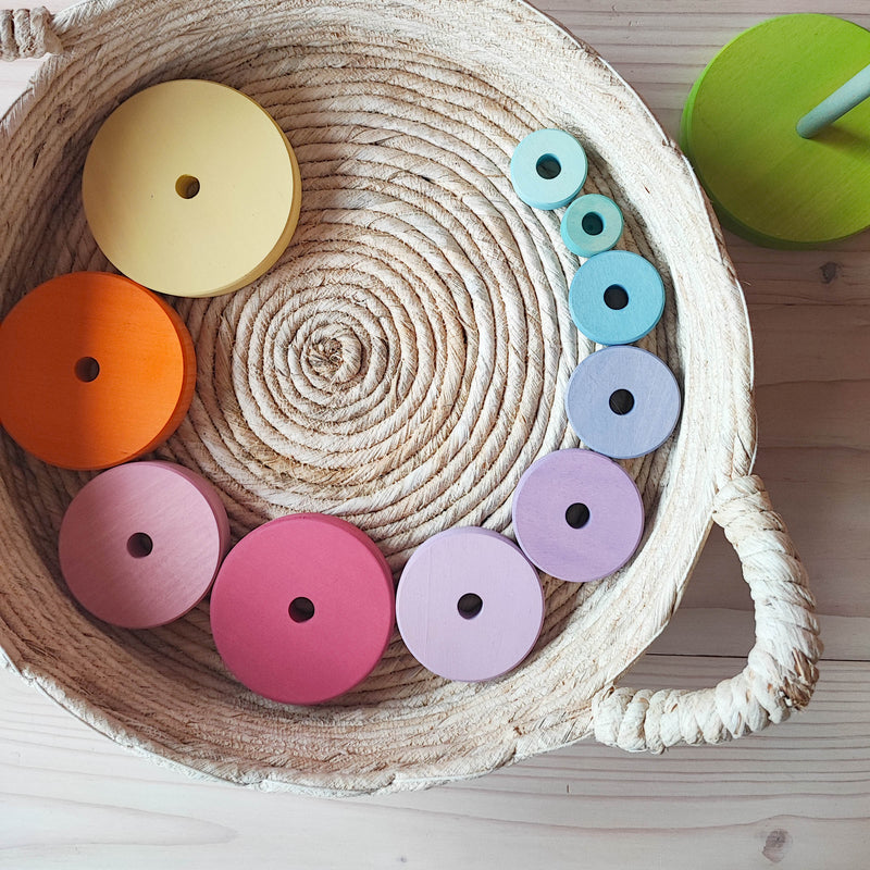 Zehn Scheiben des Grimms Scheibenturm in Pastell liegen kreisförmig am Rand in einem hellen flachen Korb. die Scheiben beginnen links oben mit der groessten in pastellgelb, und werden bis rechts oben immer kleiner. 