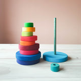 Man sieht eine hellblau lasierte runde Platte, ca 15 cm im Durchmesser. Auf ihr iste eine tuerrkisblaue Holzstange in der Mitte befestigt. Daneben sind verschieden bunte runde Holzscheiben in Regenbogenfarben aufeinander gestapelt. Die kleinste Scheib liegt davor. Man sieht das Loch in der Mitte der Scheibe.