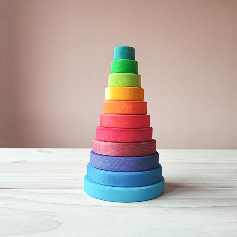 man sieht den bunten Scheibenturm von Grimms. Ein runder Stapelturm in Regenbogenfarben von helblau, mittelblau, blaulila, weinrot, rot, dunkelorange, orange, gelb, hellgruen, gruen bis tuerkisblau. Von gross bis klein.