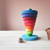 Eine Ansicht von vorne auf den Scheibenturm von Grimms. Hier sieht man die Scheiben in verkehrger Reihenfolge gestapelt. Unten ist eine runde hellblau lasierte Holzscheibe auf der vermutlich ein Stab befestigt ist. Die einzelen bunten Holzscheiben in Regenbogenfarben sind von klein nach gross auftsteigen gestapelt. 