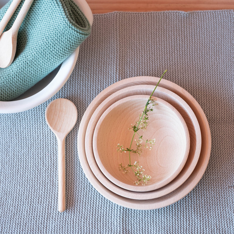 Ein Draufblick auf ein blaues Stricktuch. Darauf stehen ineinander 3 verschieden große Holzschalen mit einer Blüte darauf. Daneben liegt ein kleiner Kochlöffel für Kinder aus Holz.