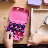 Eine Kinderhand legt ein lila Holzei zum Spielen in einen pinken Eierkarton, der schon gut mit kleinen Holzeiern gefüllt ist.