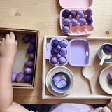Zwei Eierkarton, lila und rosa, sind mit kleinen lila Holzeiern befüllt, ein Kind greift in eine Schachtel mit mehr lila Holzeiern, es spielt Puppenküche.