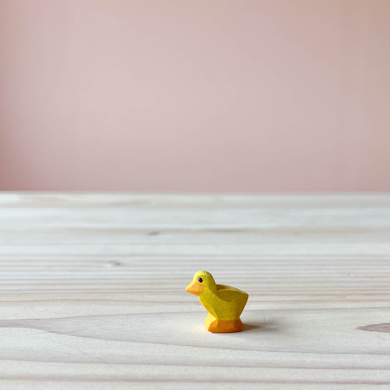 Die Ostheimer Küken Holzfigur zum Spielen ist gelb, steht auf einer Holzplatte und schaut nach links.