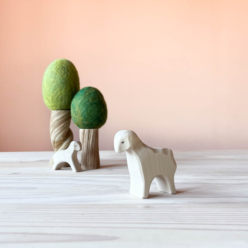 Im Vordergrund steht die Spielfigur Schaf von Ostheimer. Ein weißes Schaf, das seitlich ausgerichtet ist. Im Hintergrund zwei Filzbäume und davor steht ein weißes Lamm von Ostheimer.