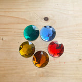 Fünf Riesenfunkelsteine in den fünf verschiedenen Farben rot, orange, gelb, grün und und blau liegen im Kreis auf einem Holzboden.