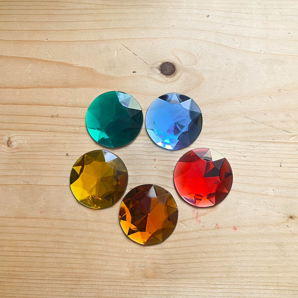 Fünf Riesenfunkelsteine in den fünf verschiedenen Farben rot, orange, gelb, grün und und blau liegen im Kreis auf einem Holzboden.