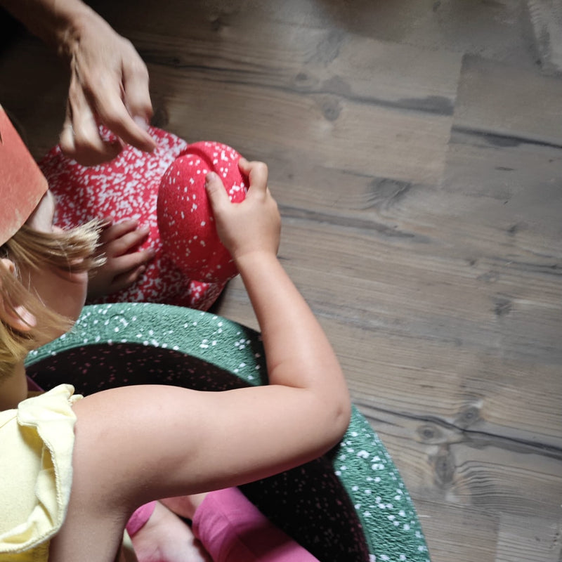 Ein Kind in einem grünen Grow Stapelstein spielt mit zwei rot-weiß gesprenkelten Original- und Mini-Stapelsteinen vor sich, alle Teil des Limited Stapelstein®Blush Special Triology Set.