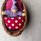 Das Wichtelnest von Kokomoo: ein Weidenkörbchen als Puppenbett, in dem ein lila Nanchen Wichtel in einer rot gemusterten Bettwäsche kuschelt. 