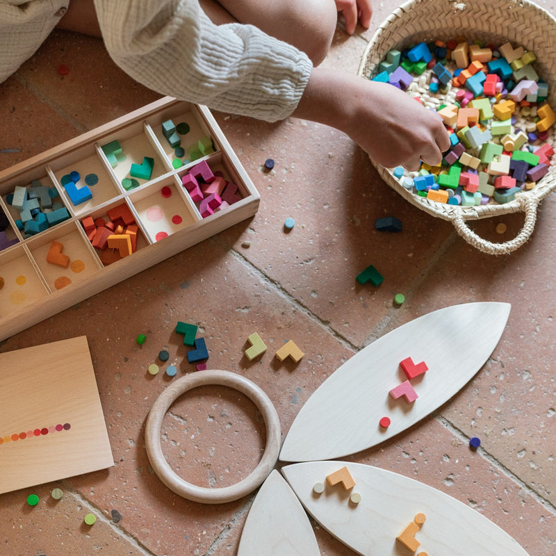 Eine Kinderhand, die mit den Mini-Holzsteinen von Grapat Mis, Match and Bloom  spielt, sie aus einem Korb nimmt und sortiert. Daneben liegen blütenförmige Holzscheiben und Holzringe am Boden.