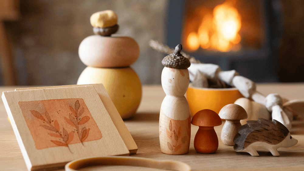 Das Spiele- und Forscherset für Kinder "Hello Autumn" von Grapat und Ostheimer steht auf einem Tisch vor dem Kamin. Der Igel, die beiden Holzpilze, die Nins Spielfigur und die kleine Blätterpresse.