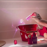 Ein roter kleiner Eimer aus Emaille, ein paar rote Holzfiguren und eine Hand, die ins Bild hineinreicht und einen rosafarbenen Acrylquader der Firma Bauspiel in der Hand hält und ihn dreht. Durch den Lichteinfall wirft der transparente Bauklotz wunderschöne rosastrahlende Muster an die dahinterliegende Wand.