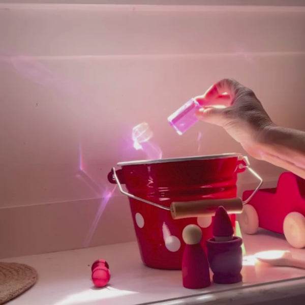 Ein roter kleiner Eimer aus Emaille, ein paar rote Holzfiguren und eine Hand, die ins Bild hineinreicht und einen rosafarbenen Acrylquader der Firma Bauspiel in der Hand hält und ihn dreht. Durch den Lichteinfall wirft der transparente Bauklotz wunderschöne rosastrahlende Muster an die dahinterliegende Wand.
