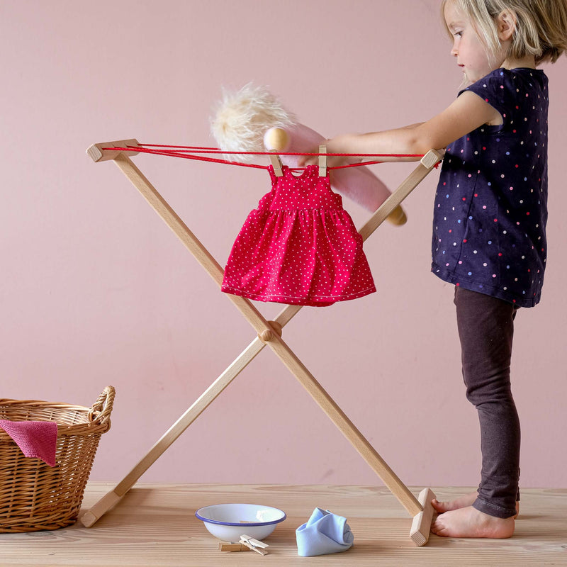 Ein Kind steht neben dem Wäscheständer von Glückskäfer und hält eine Puppe. Das rote Puppenkleid hängt gerade zum Trocknen an der Wäscheleine.