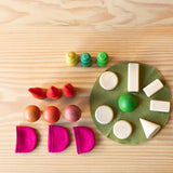 Draufsicht auf die einzelen Bestandteile des Spielesets Freunde im Wald. Auf einem gruenen Filzkreis liegen geometrische Formen - kegel, Quarder Scheibenin naturhoz und eine gruene Halkugel. Daneben stehen 3 handbemalte Holzpilze, drei gruene und 2 rote Holzkegel und es liegen 3 pinkfarbene Filztaeschen davor.