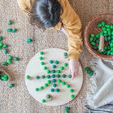 Ein Kind liegt am Boden, es legt ein Muster mit verschieden geformten gruenen Mandala Steinen auf die große, runde Holzplatte von Grapat.