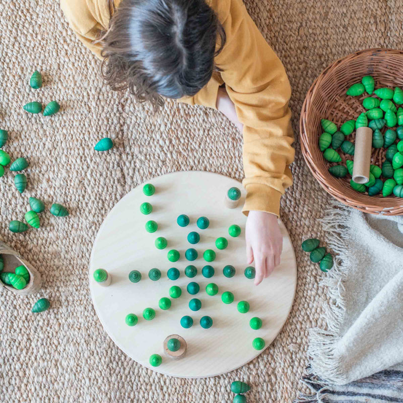 Ein Kind liegt am Boden, es legt ein Muster mit verschieden geformten gruenen Mandala Steinen auf die große, runde Holzplatte von Grapat.