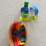 Blick von oben. Man sieht im bild links unten eine ovalen Waeschekorb aus Weidenholz. Darin liegen einzeln die bunten verschieden grossen Boegen des Grimms regenbogens. Oberhalb stehen und liegen 4 Regenbogen elemente in Blau und Gruentoenen.