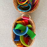 Blick on oben auf 2 ovale Weidenkoerbe mit Henkel. im vorderen liegen alle teile des Grimms Regenbogens in 12 Farben ganz durcheinandergemischt. Im Bild oben - auch ein Korb mit bunten runden Motorikringen der Firma gonge.