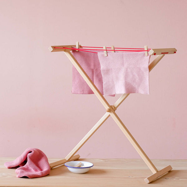 Der kleine Wäscheständer für Kinder aus Holz, es sind zwei rosa Stricktücher aufgehängt, am Boden steht eine Emailleschüssel mit Wäscheklammern, daneben liegt ein pinkes Tuch.