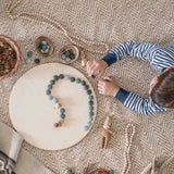 Ein Kind liegt am Boden, es legt Muster mit grauen Mandala Steinen auf die große, runde Holzplatte von Grapat.