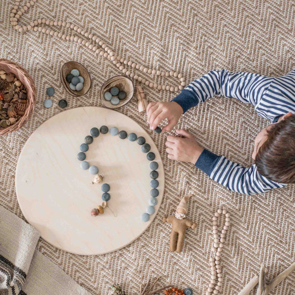 Ein Kind liegt am Boden, es legt Muster mit grauen Mandala Steinen auf die große, runde Holzplatte von Grapat.